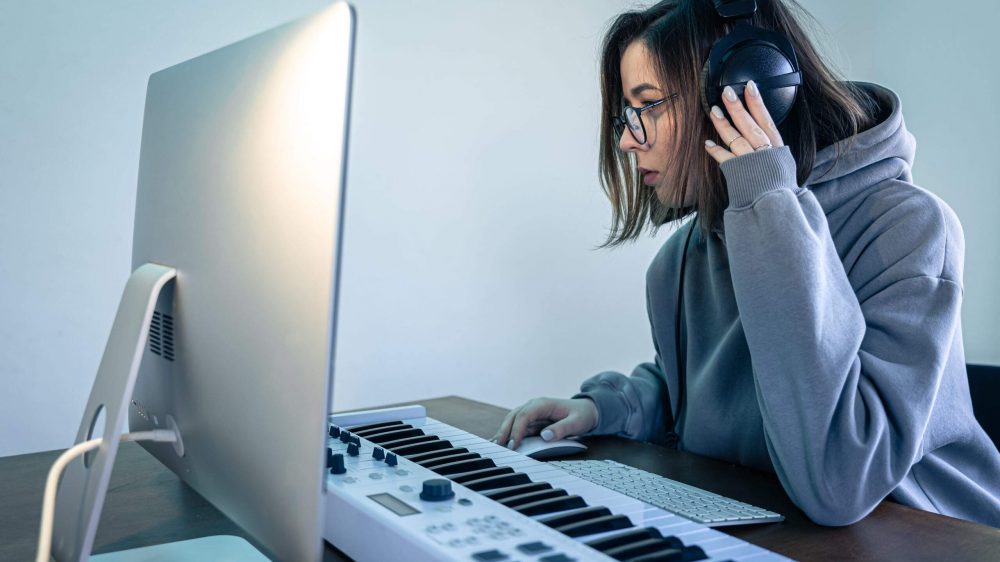 A young woman musician at workplace, creates music using musical keyboard and computer, freelance, musical creativity.
